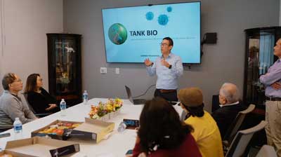 Man giving a presentation in front of a TV screen in an office with students around the table<br />
